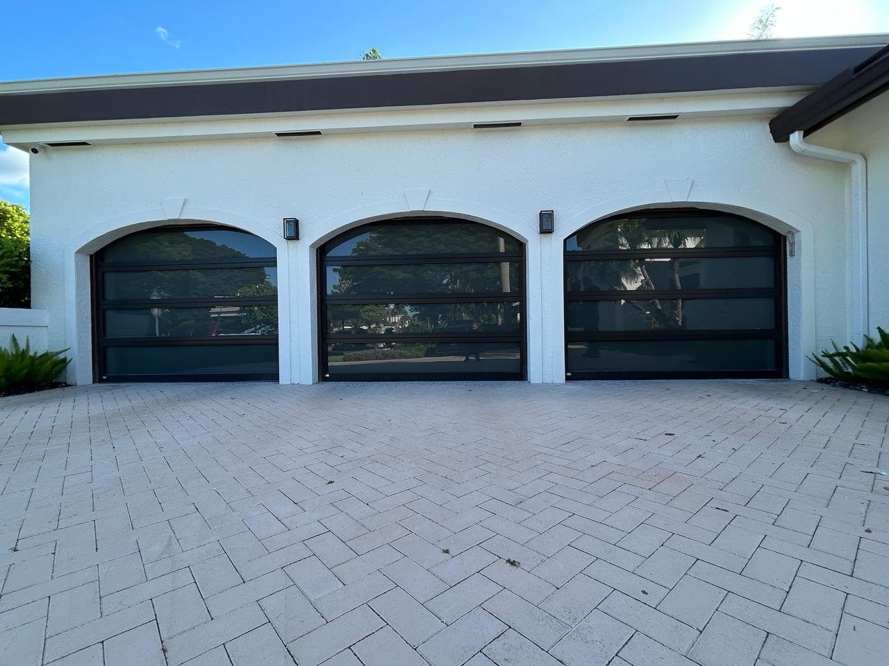 Three arched black-frame impact glass AA Impact garage doors on a white South Florida home with a herringbone paver driveway under a clear blue sky.