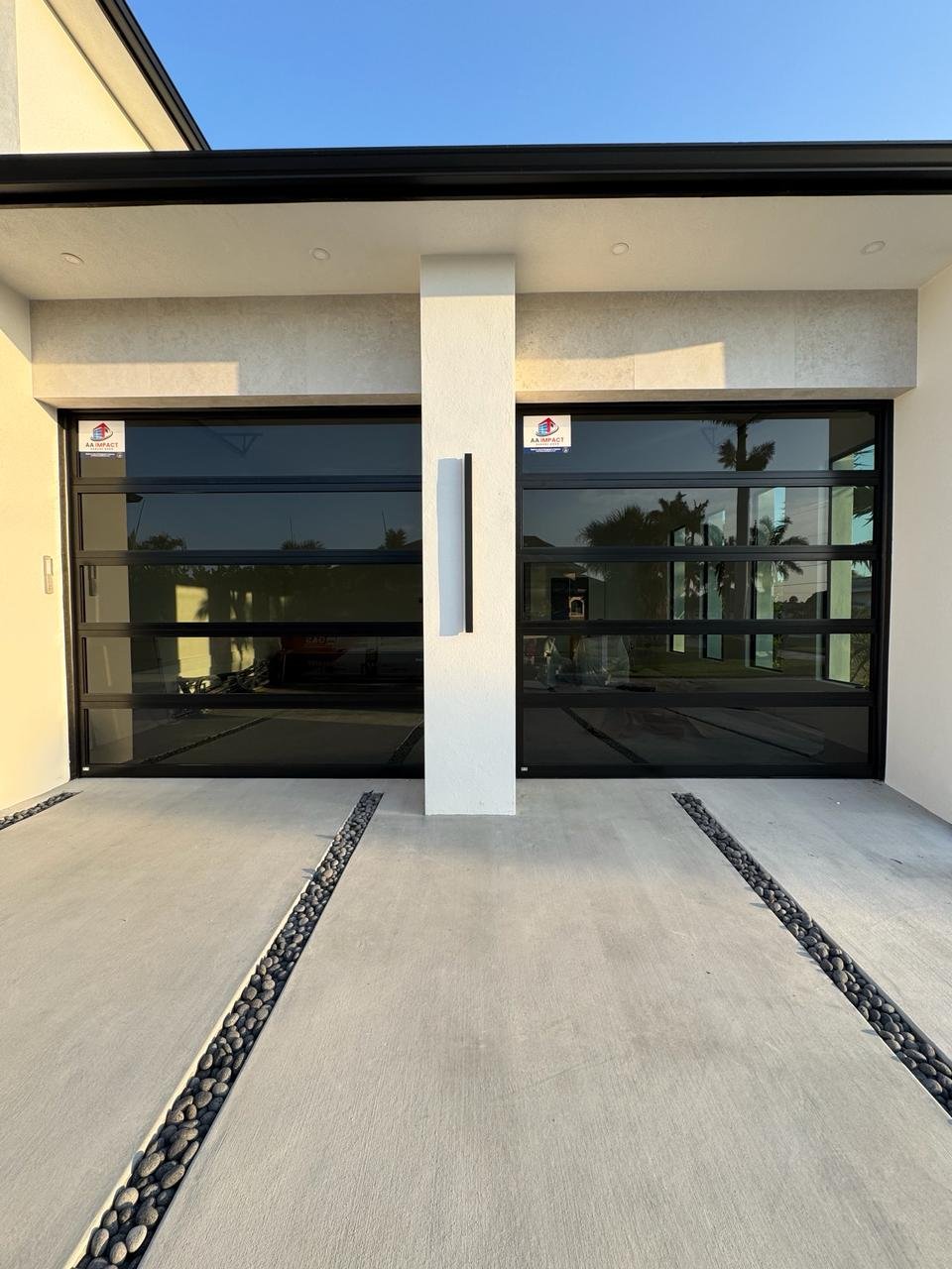 AA Impact Series 1200 full-view impact glass — twin black-frame doors flanking a white column on a modern South Florida home.