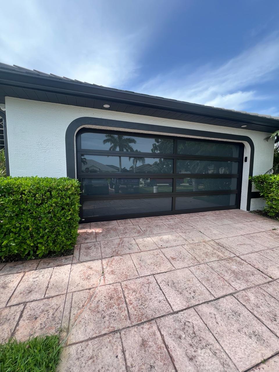 A single black-frame full-view impact glass AA Impact garage door with a curved surround on a white South Florida home, framed by a manicured hedge and brick pavers.