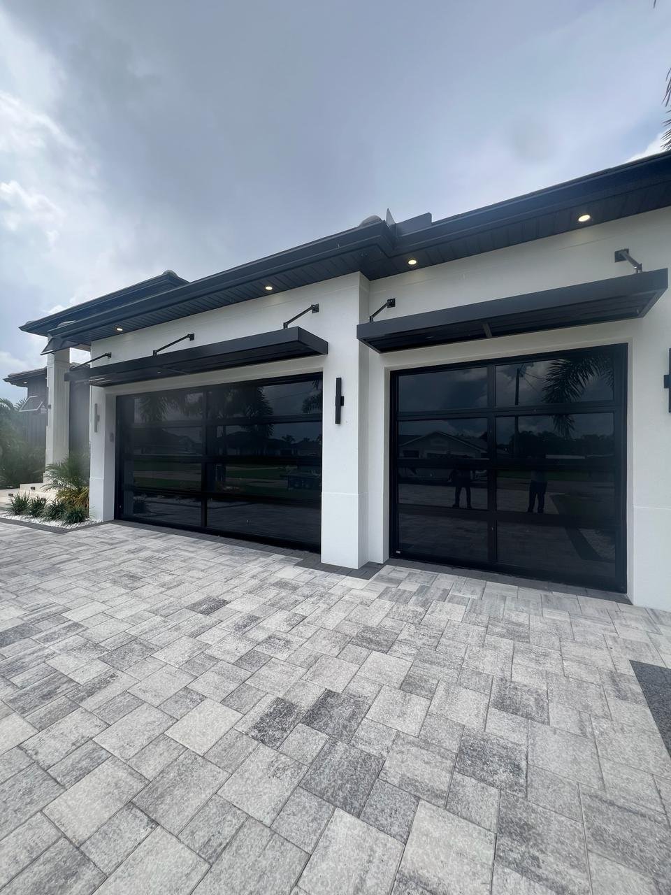 Two black-frame full-view impact glass AA Impact garage doors on a white South Florida home with a light stone paver driveway under an overcast sky.