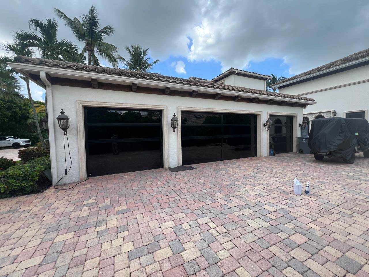 A three-bay AA Impact black-frame impact glass garage door on a tile-roof South Florida estate with palm trees and a red-toned paver driveway.
