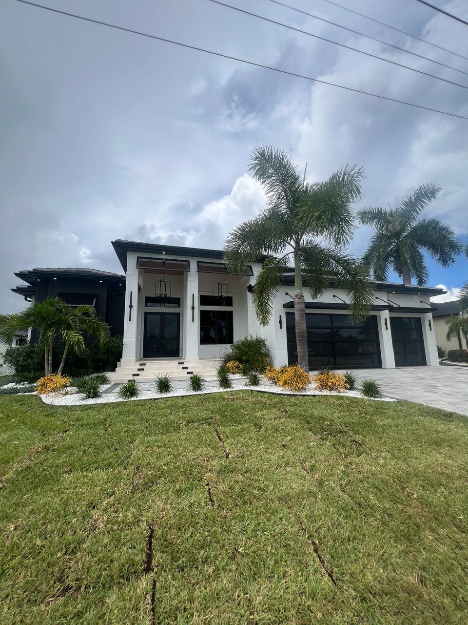 Full front elevation of a modern white South Florida home with black-frame impact glass AA Impact garage doors, entry portico, and palm-lined landscaping.