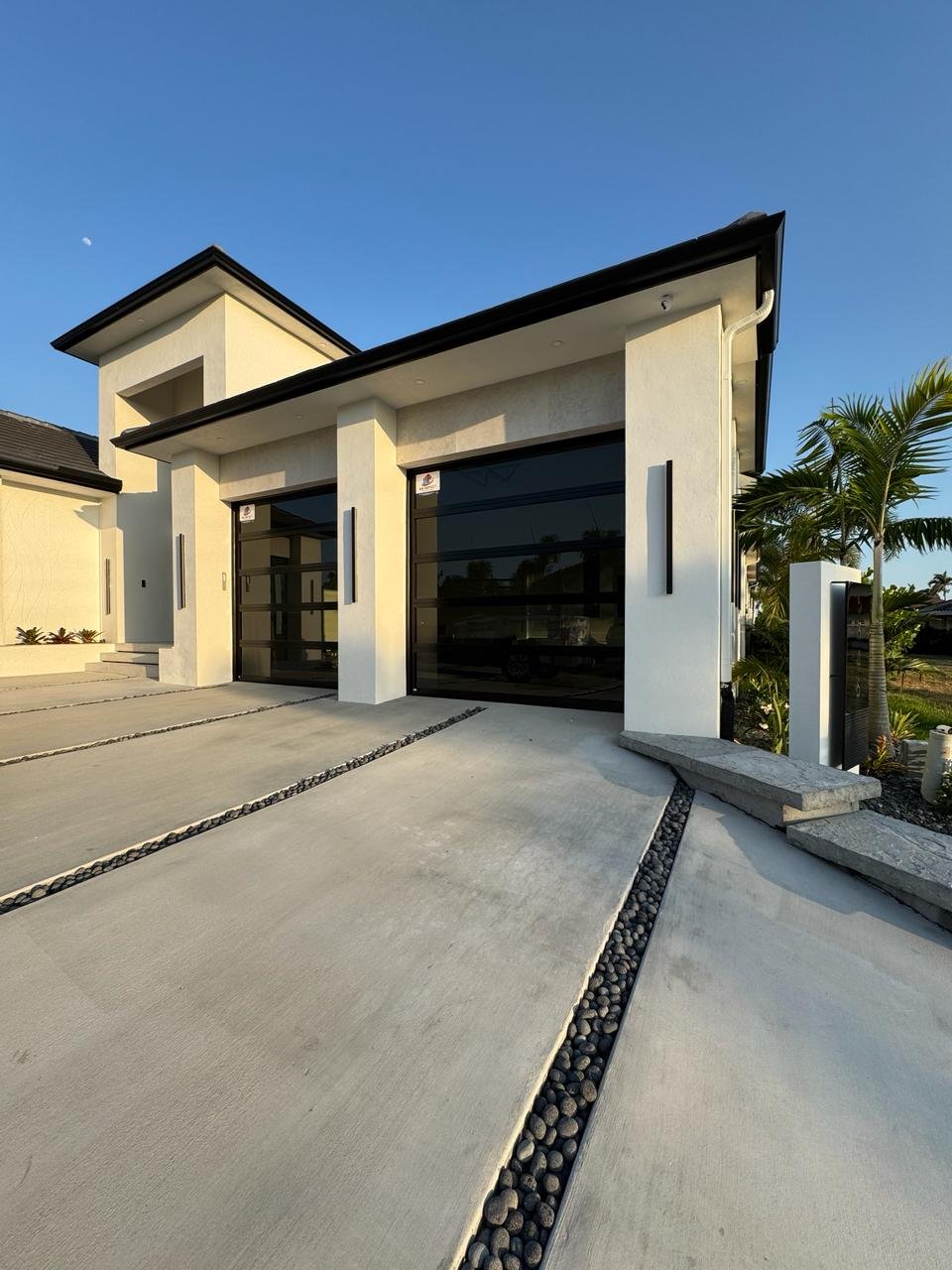 Two black-frame full-view impact glass AA Impact garage doors on a modern white South Florida home at golden hour, with a poured-concrete driveway and river-rock inlays.