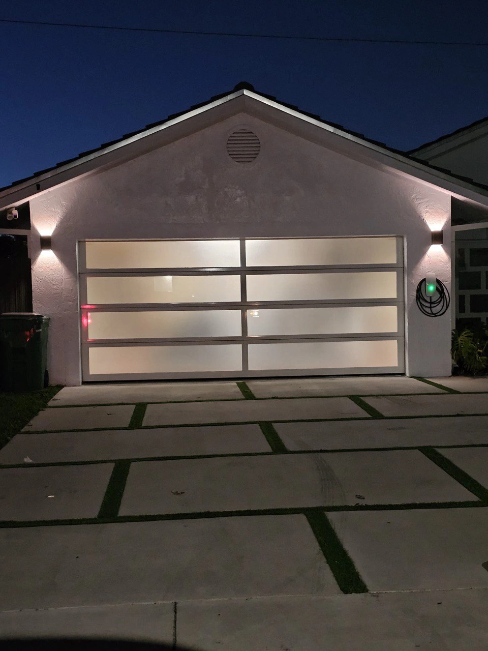 A frosted white-glass AA Impact impact garage door lit from within at night on a South Florida home, with wall-washer sconces and a ribbon driveway.