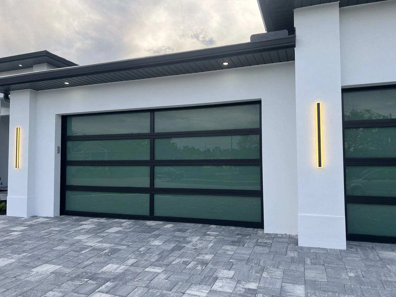 AA Impact Series 1600 impact glass garage door with green-tinted full-view glass and a black frame on a modern South Florida home at dusk.
